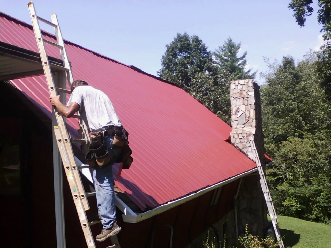 Roofer on a ladder beside a red metal roof and stone chimney — Soffit & Fascia Repair by Roofing Brothers in Azle